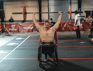 Mike Egan - Marine Wounded In Battle Competing at the 2023 WheelWOD Games, sitting in his wheelchair holding a barbell over his head with 95 pounds for reps.