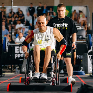 Man in a wheelchair participating in a CrossFit event with another person assisting.