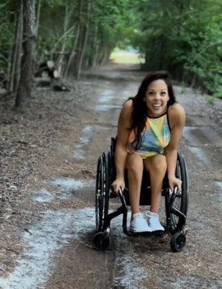 Liz Mozingo in her wheelchair on a gravel path with trees overhanging and appears to have rained recently. she is kind of dipping down to the camera and smiling really cute