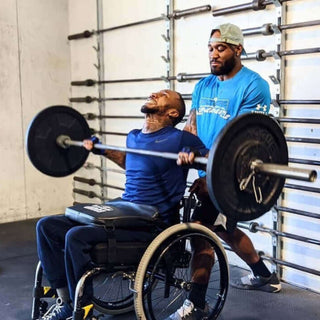 A Man in a wheelchair using a LapMat to protect his legs while using a barbell in a gym.