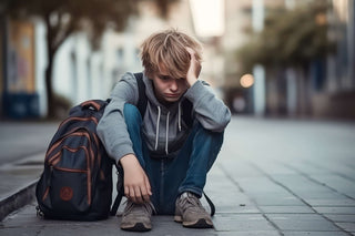 A picture of a young man sitting on the ground with his backpack next to him, head in hand, looking dejected.