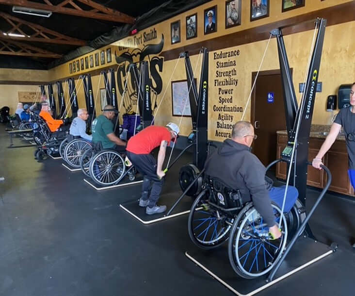Several Men in wheelchairs using the adaptive wider base and Concept2 Ski Erg in a gym setting.