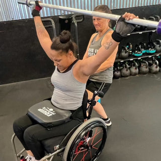 Wheelchair athlete holding a barbell over his her head with a handler behind her