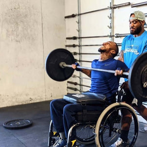 A Man in a wheelchair using a LapMat to protect his legs while using a barbell in a gym.